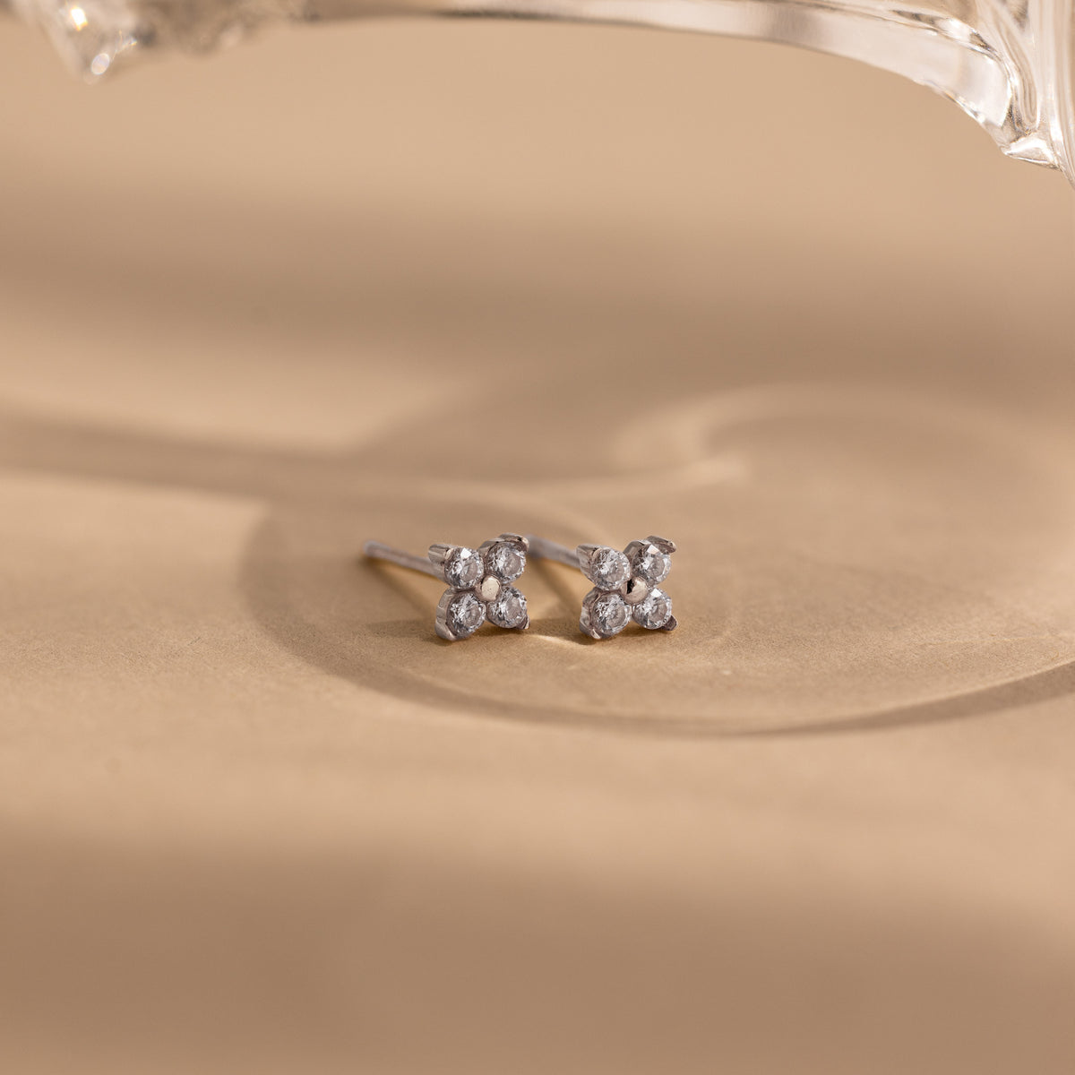 A pair of small silver stud earrings with four clear stones arranged in a flower shape, resting on a beige surface with a soft shadow and a blurred clear object at the top.