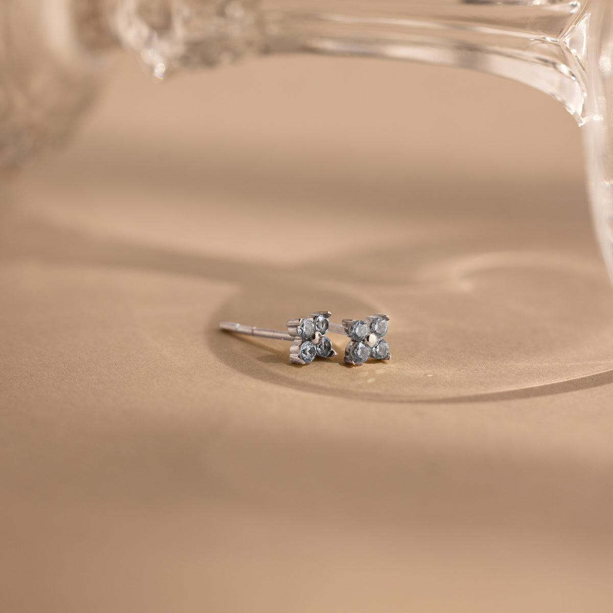 A pair of small silver stud earrings with four light blue stones arranged in a flower shape, resting on a warm beige surface with a softly blurred glass object in the background.