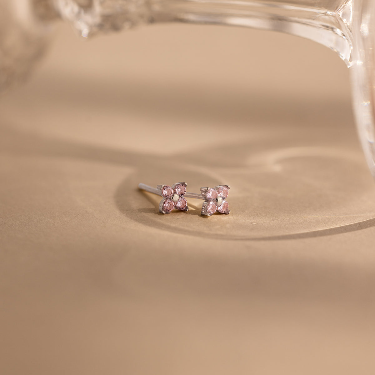 A pair of small stud earrings with light pink gemstones in a bow-like cluster setting, photographed on a warm beige surface with a soft-focus glass object in the background.