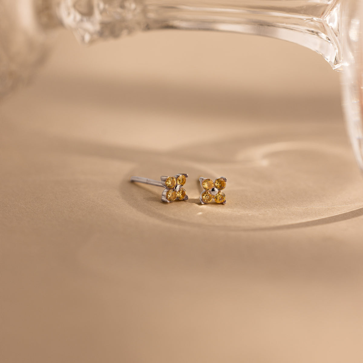 A pair of small silver stud earrings with four round yellow gemstones, resting on a warm beige surface with a soft-focus glass object in the background.