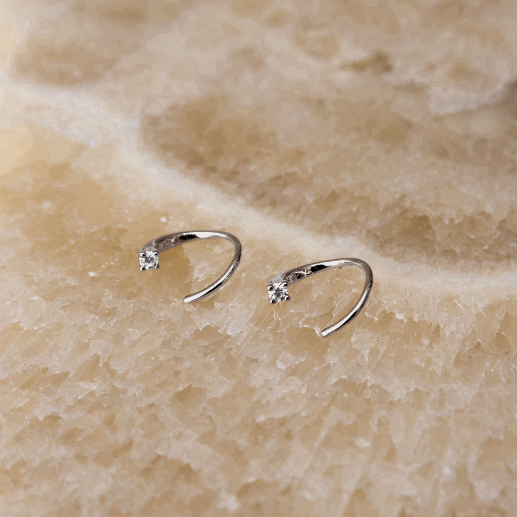 A pair of small silver open hoop earrings, each set with a single clear gemstone, displayed on a textured beige stone surface.