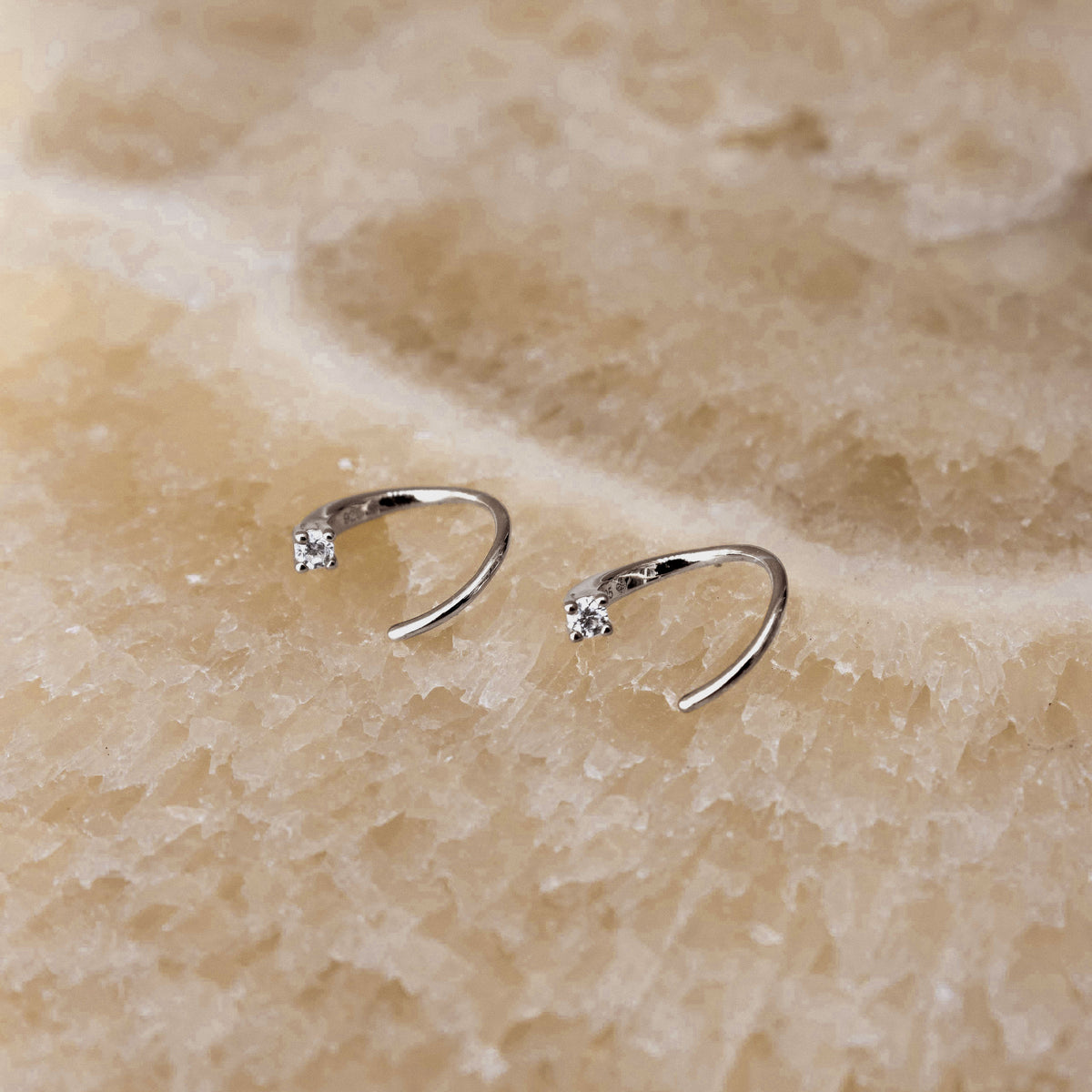 A pair of small silver open hoop earrings, each set with a single clear gemstone, displayed on a textured beige stone surface.