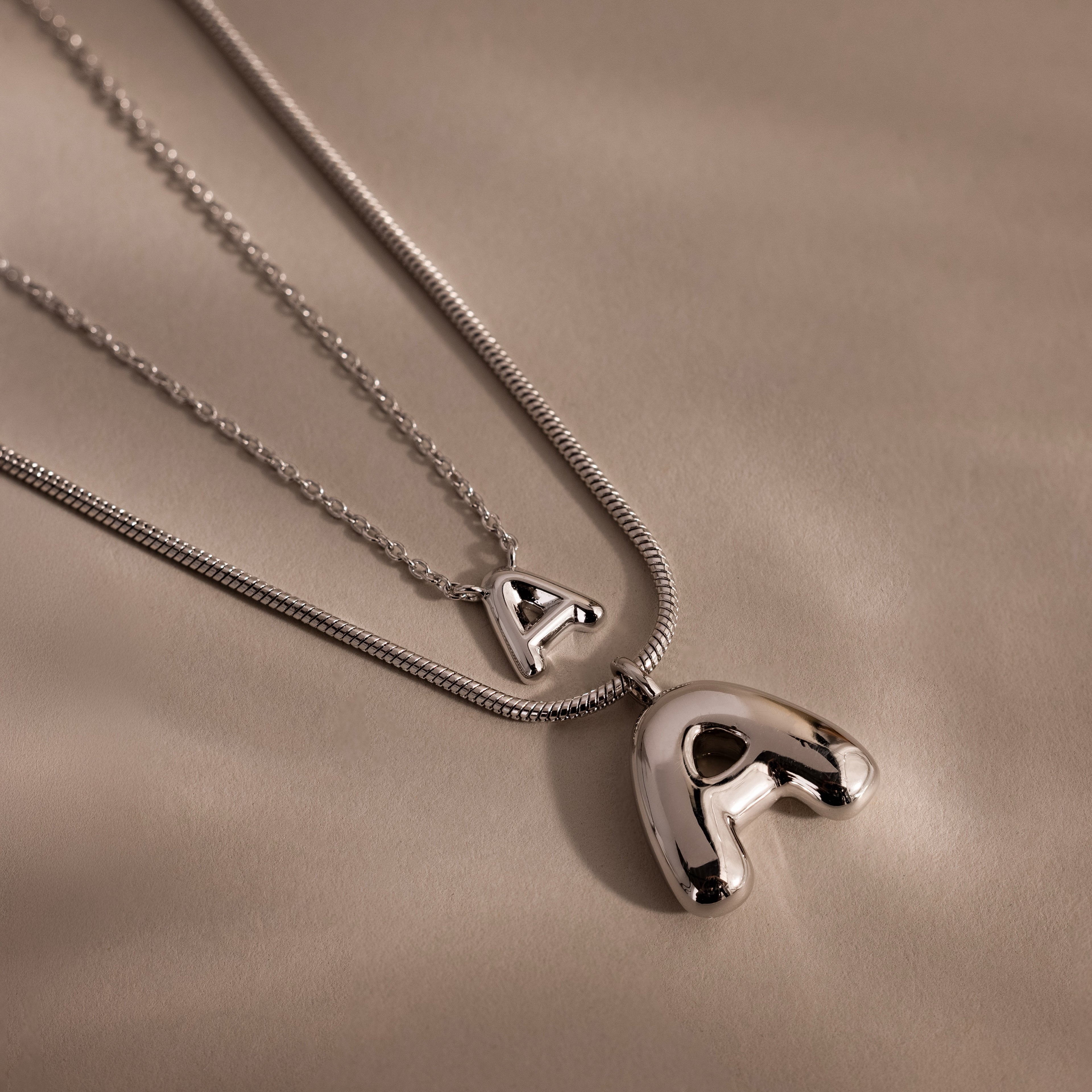 Two Bubble Letter Necklaces in White Gold with shiny letter A pendants rest on a smooth beige surface—ideal for personalized jewelry lovers or anyone seeking a classic Bubble Letter Necklace style.
