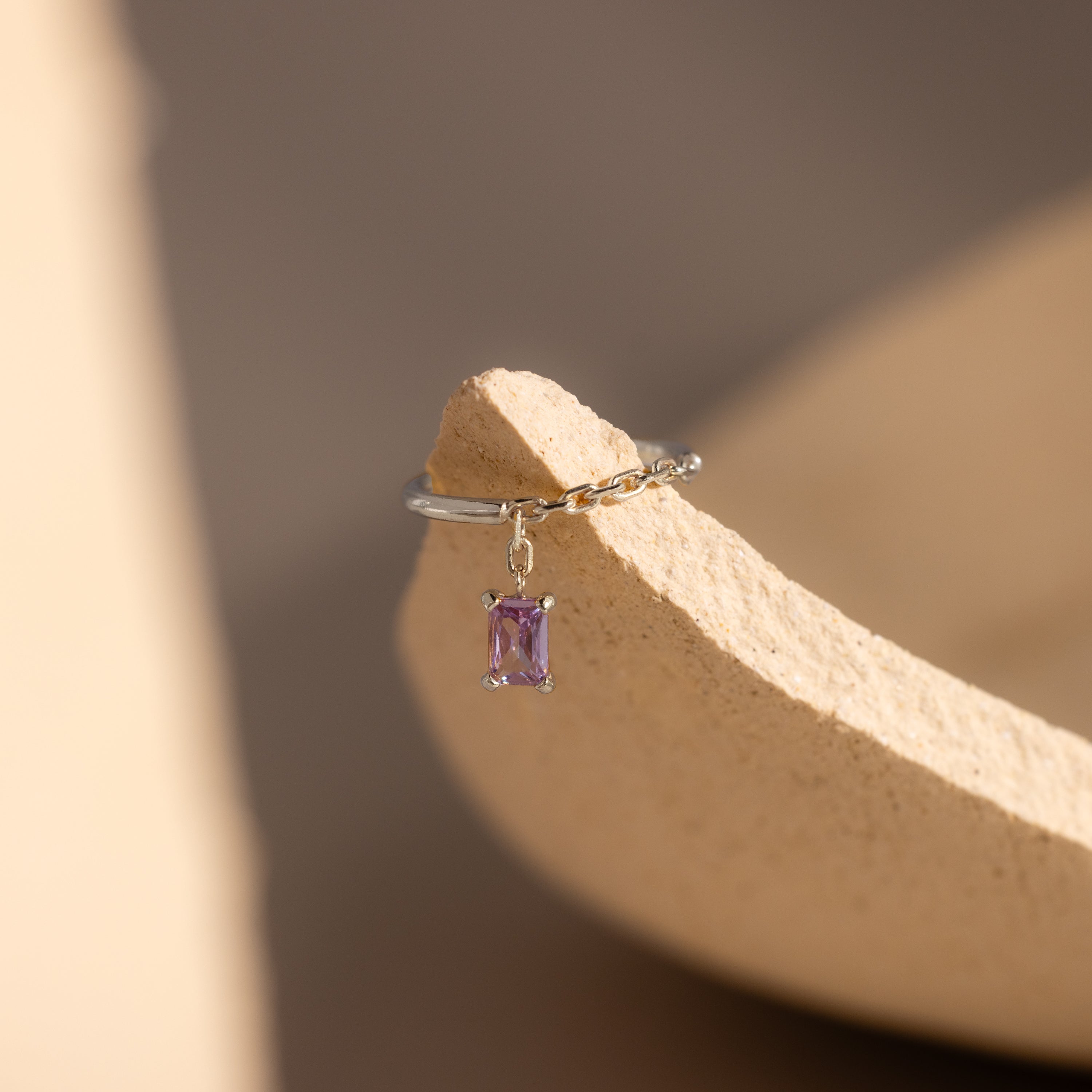 The Alexandrite Drop Chain Ring, featuring a silver band with a dangling rectangular purple gemstone in an emerald cut, is displayed on a textured beige surface.
