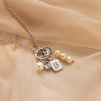 A silver chain necklace with a charm set featuring three white pearls, a small silver bag charm, a square silver block charm engraved with the letter 'B', and a tiny silver bead, displayed on a draped beige fabric background.
