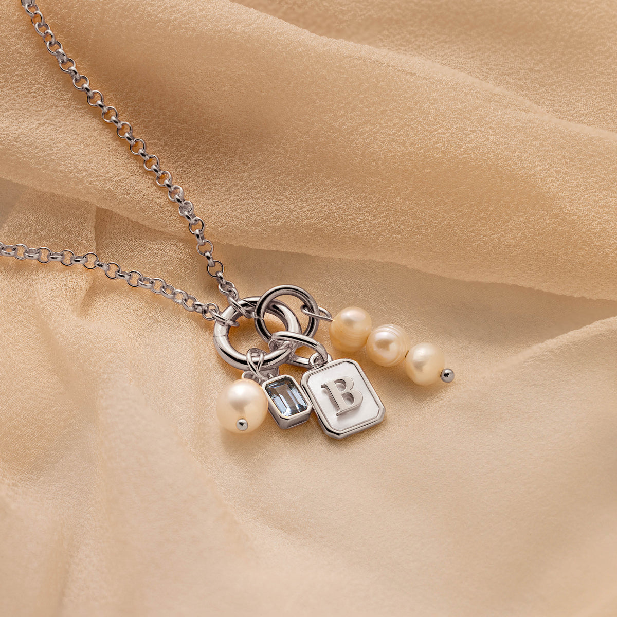 A silver chain necklace with a charm set featuring three white pearls, a small silver bag charm, a square silver block charm engraved with the letter 'B', and a tiny silver bead, displayed on a draped beige fabric background.