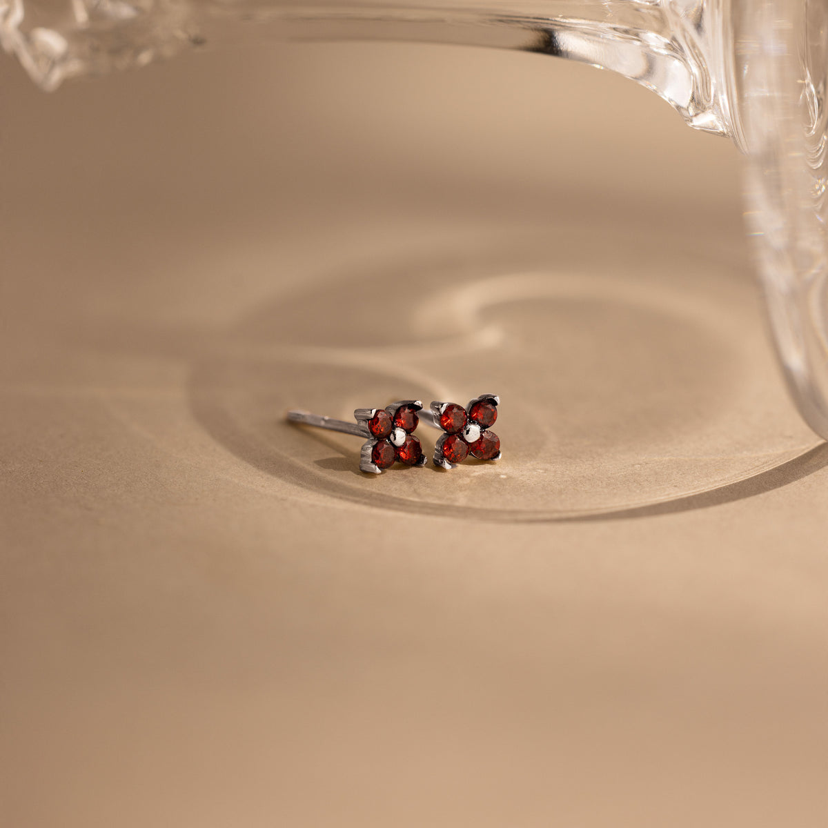 A pair of silver stud earrings with four small red gemstones arranged in a square, resting on a beige surface with a soft shadow and a glass object partially visible at the top.