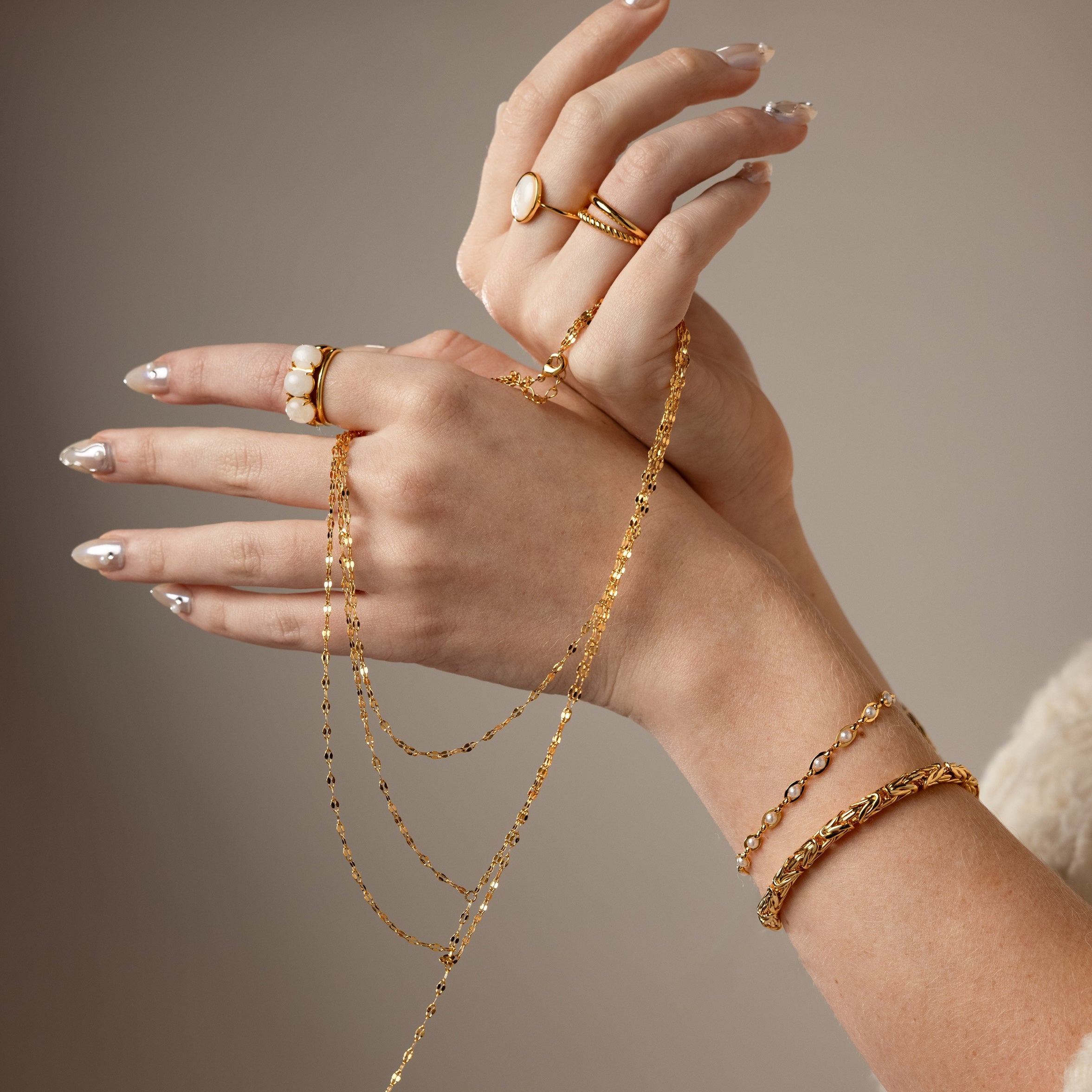 Two hands with manicured nails wearing gold rings and bracelets hold the Sparkling Layered Lariat Necklace in 18K Gold, perfect for effortless layering and chic summer style.