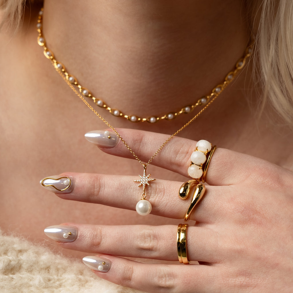 A hand with pearl-accented nails, gold rings, and layered gold necklaces featuring pearl jewelry and the North Star Pearl Necklace in 18K Gold.