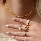 A hand with pearl-accented nails, gold rings, and layered gold necklaces featuring pearl jewelry and the North Star Pearl Necklace in 18K Gold.