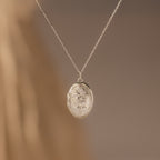 The Floral Handwriting Locket Necklace features an oval silver pendant with delicate engravings, hanging from a fine chain and set against a softly blurred neutral background.