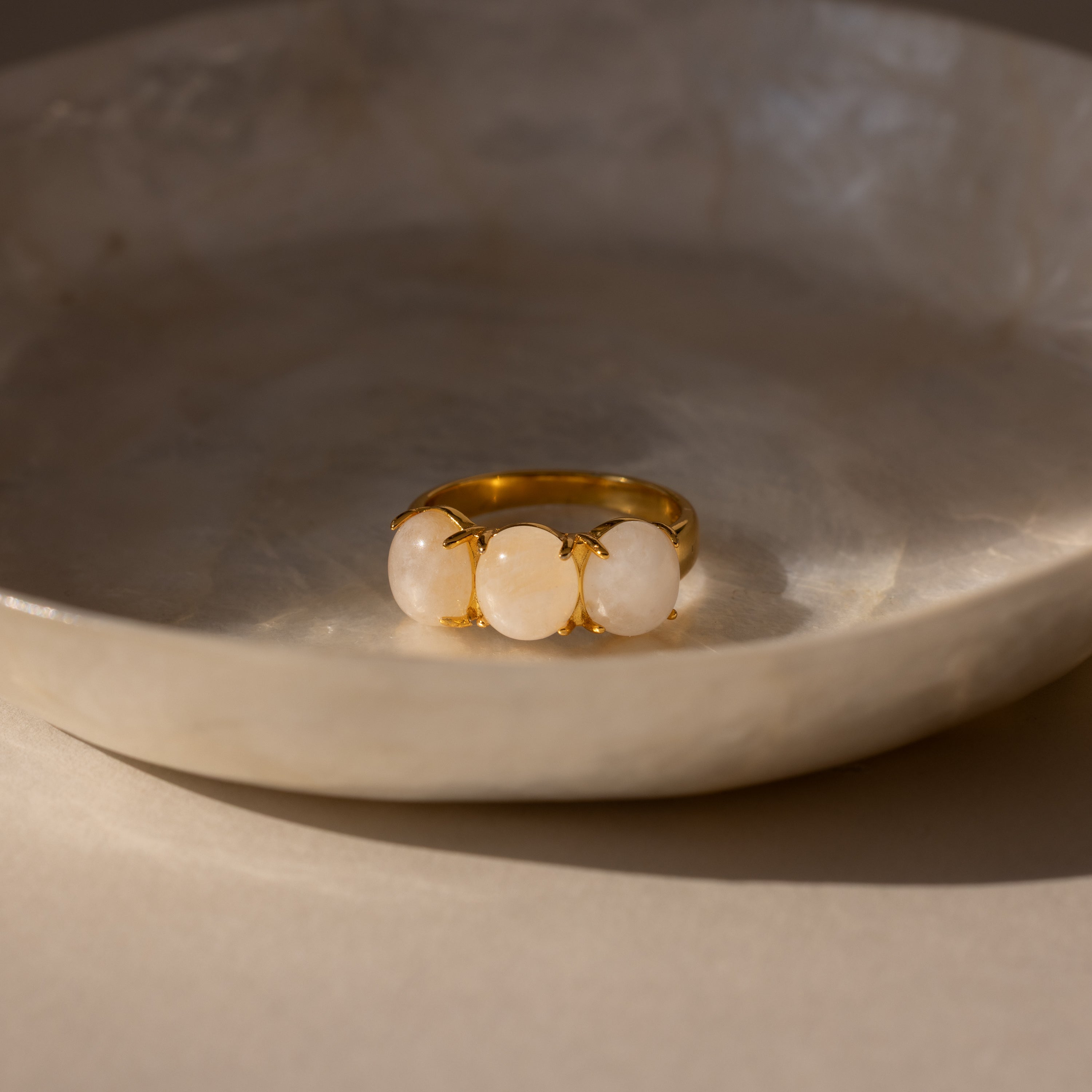 The Cosmic Moonstone Ring, featuring three round white stones, is displayed on a beige ceramic dish in soft natural light—a stunning piece of boho jewelry.