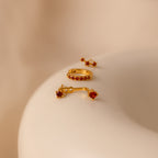 The Garnet Earring Trio Set, featuring three elegant gold earrings adorned with red gemstones, is displayed on a smooth beige surface.