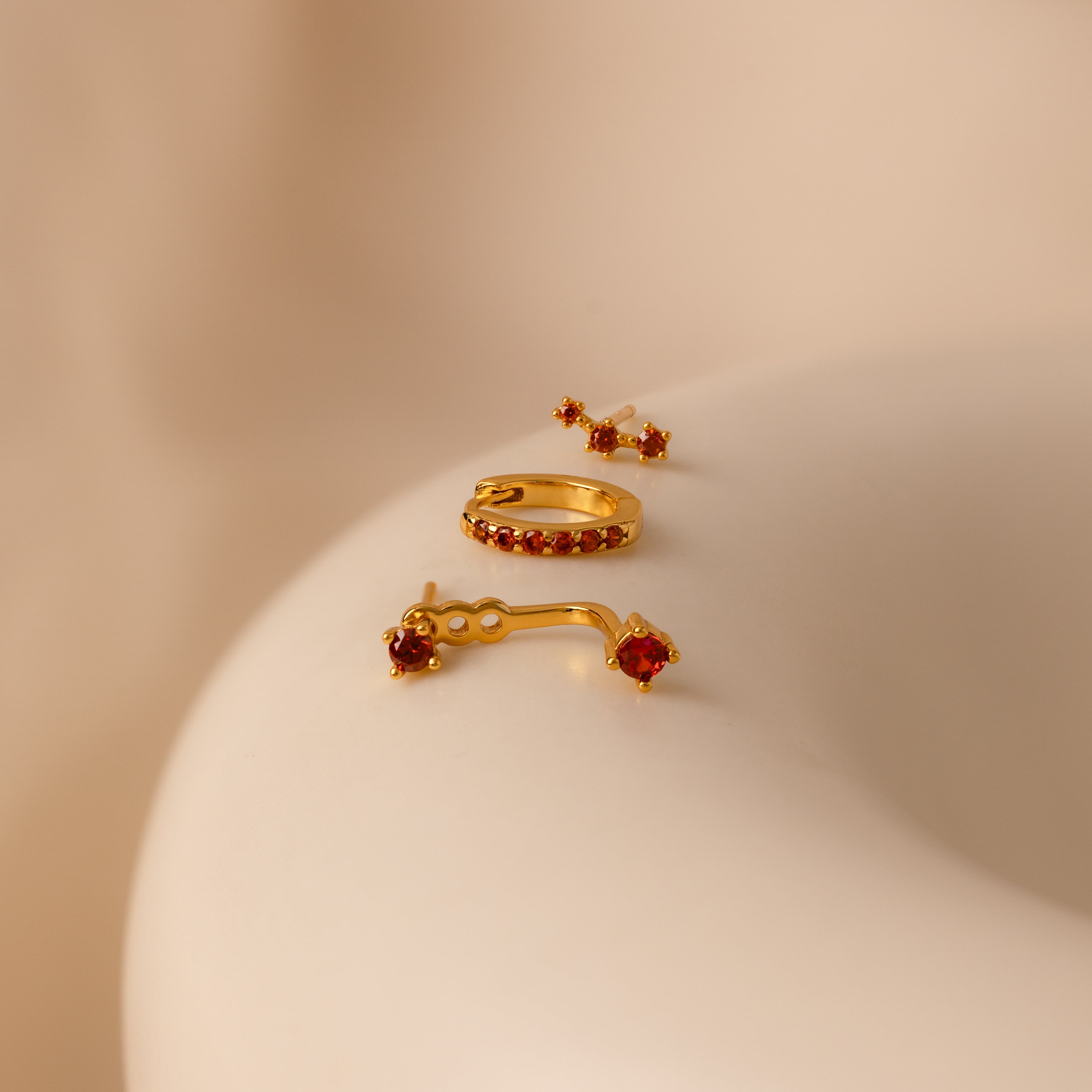 Birthstone Earring Trio Set with pavé huggie hoops and red gemstones, showcased on a smooth beige surface under soft lighting.