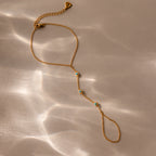 The Turquoise Hand Chain Bracelet in 18K Gold features delicate blue stones, reminiscent of a turquoise bracelet, displayed on a beige surface with soft light reflections.