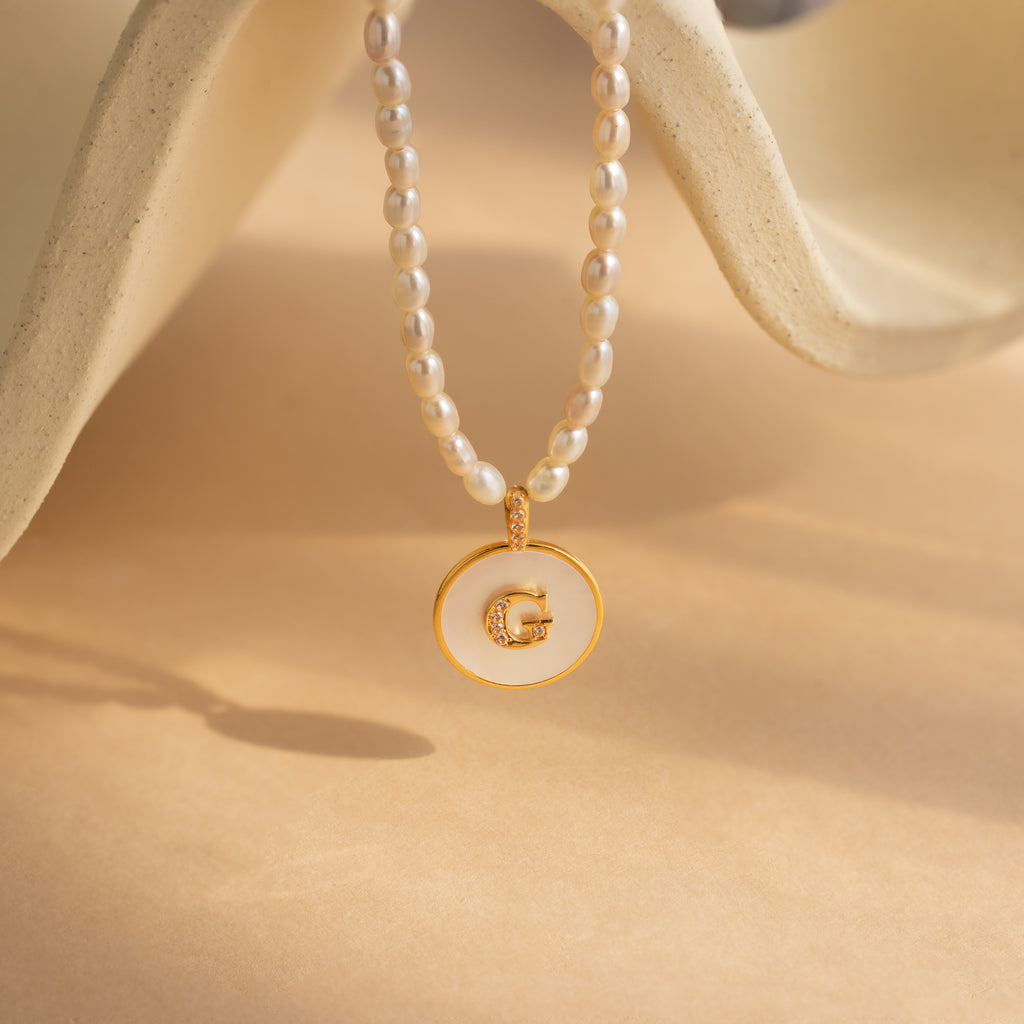 The Pearl Initial Disc Necklace features a gold and white pendant with the letter G on a beige background, offering an elegant and customizable accessory—perfect for adding personalized style.