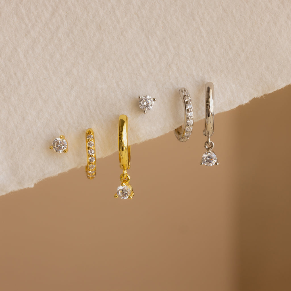 The Classic Diamond Earrings Set features six elegant gold and silver earrings with small diamonds, including classic stud styles, beautifully displayed on a textured cream surface.