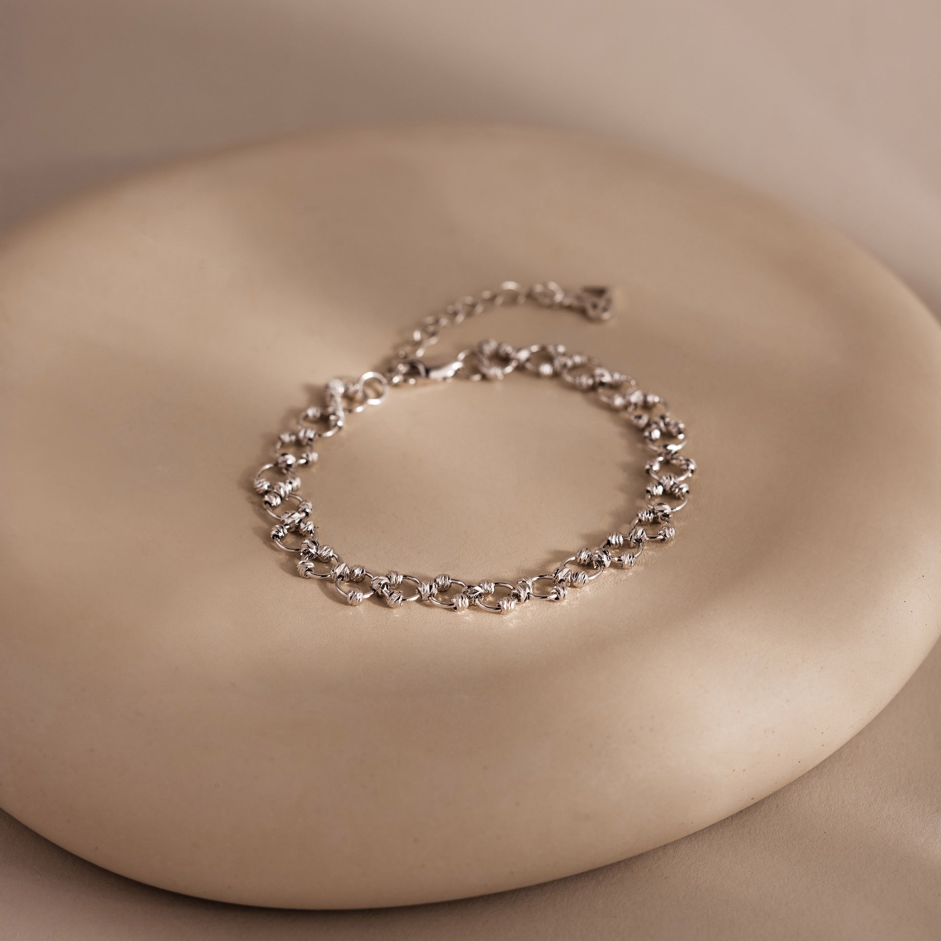 The Orbit Chain Bracelet in Silver, featuring delicate floral links, is showcased on a smooth round beige surface—an exquisite piece of elegant jewelry.