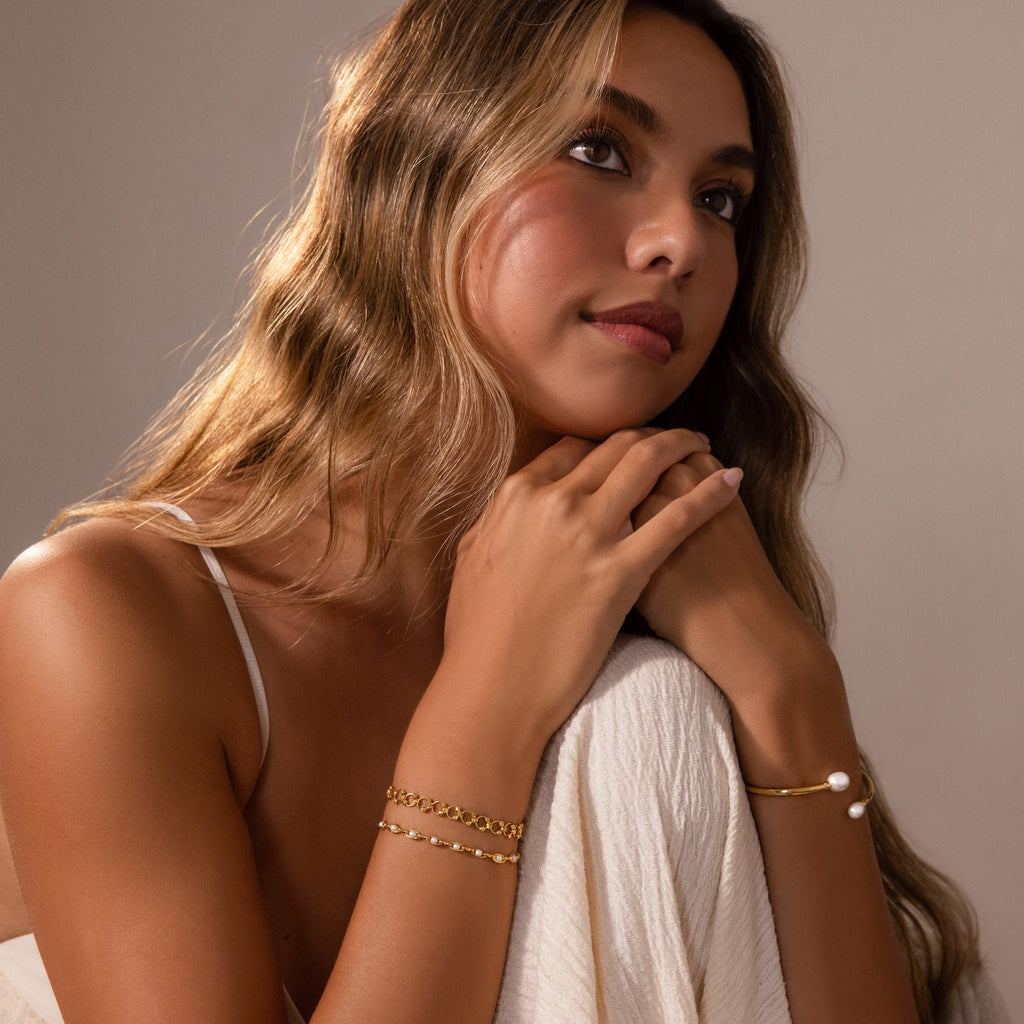 A woman with wavy hair wears the Orbit Chain Bracelet in 18K Gold and gold bracelets, resting her chin on her hands and gazing thoughtfully upward.
