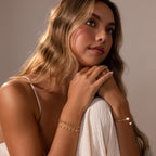 A woman with wavy hair wears the Orbit Chain Bracelet in 18K Gold and gold bracelets, resting her chin on her hands and gazing thoughtfully upward.