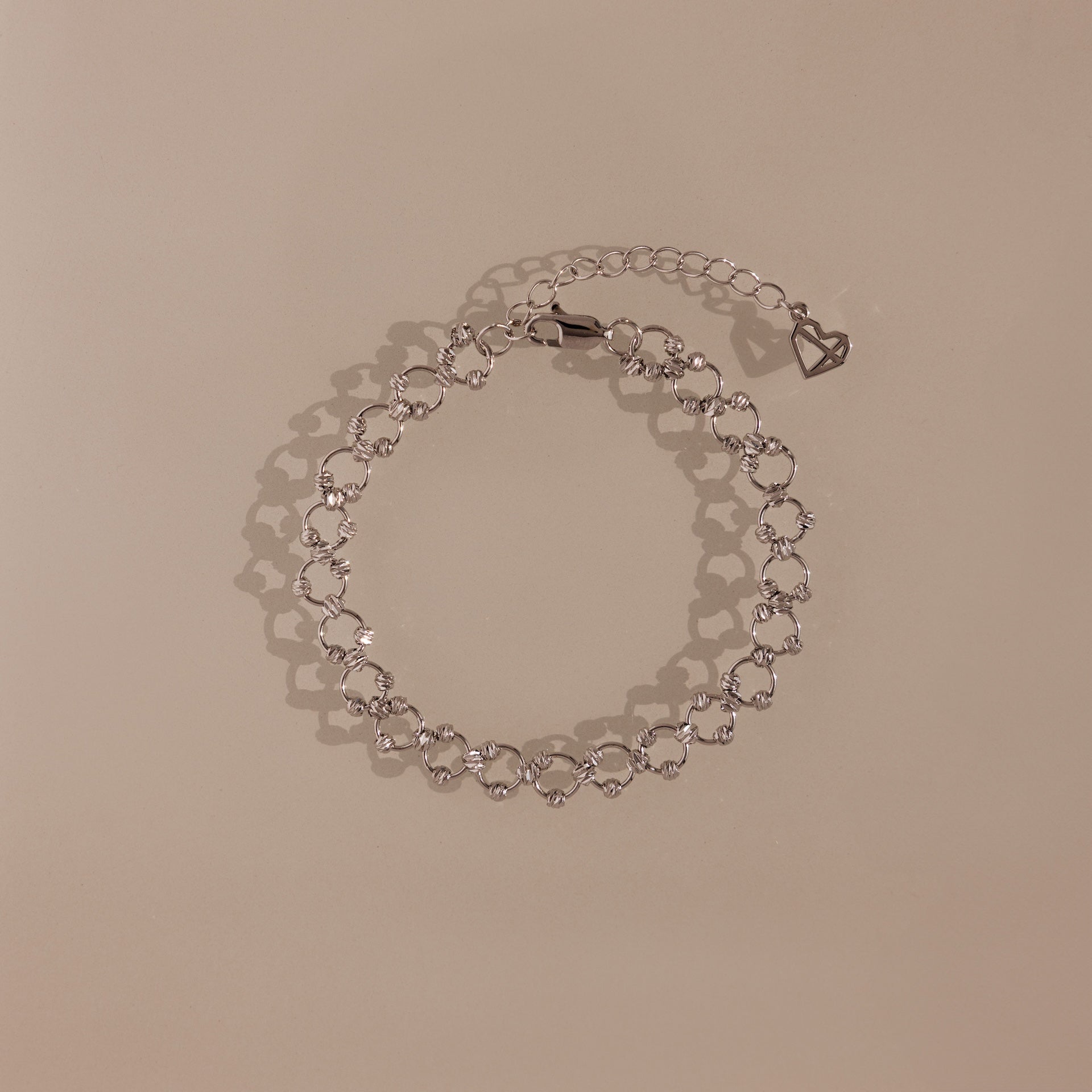 The Orbit Chain Bracelet in Silver features delicate heart-shaped links and an adjustable chain, showcased on a beige background—ideal for fans of elegant jewelry.