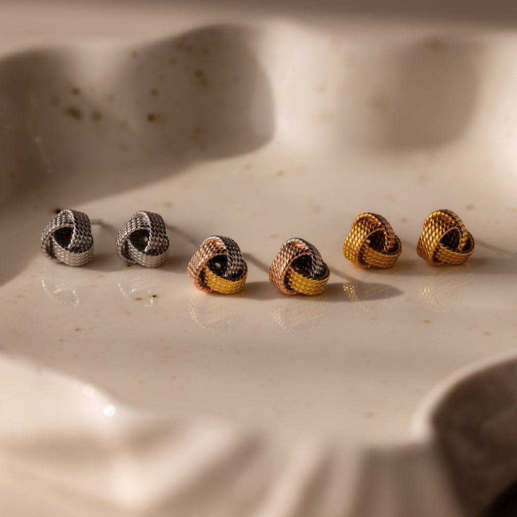 The Rope Knot Studs in Mixed Metal feature three pairs of knot-shaped earrings in silver, rose gold, and gold, elegantly arranged on a white ceramic dish—ideal for versatile everyday style.