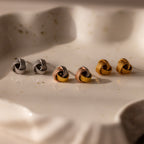 The Rope Knot Studs in Mixed Metal feature three pairs of knot-shaped earrings in silver, rose gold, and gold, elegantly arranged on a white ceramic dish—ideal for versatile everyday style.
