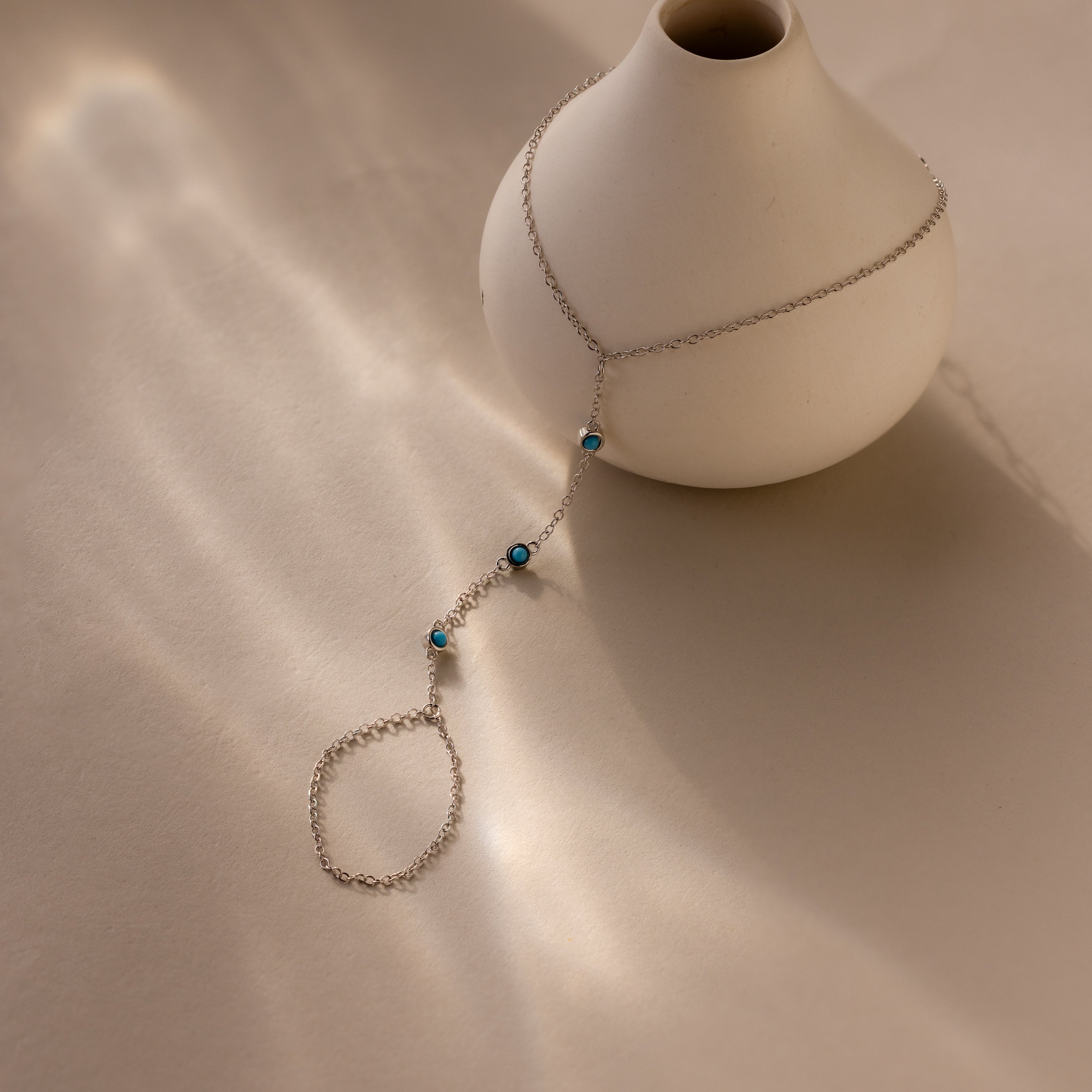 The Turquoise Hand Chain Bracelet, adorned with blue stones, is elegantly draped on a white vase against a neutral background with soft lighting.