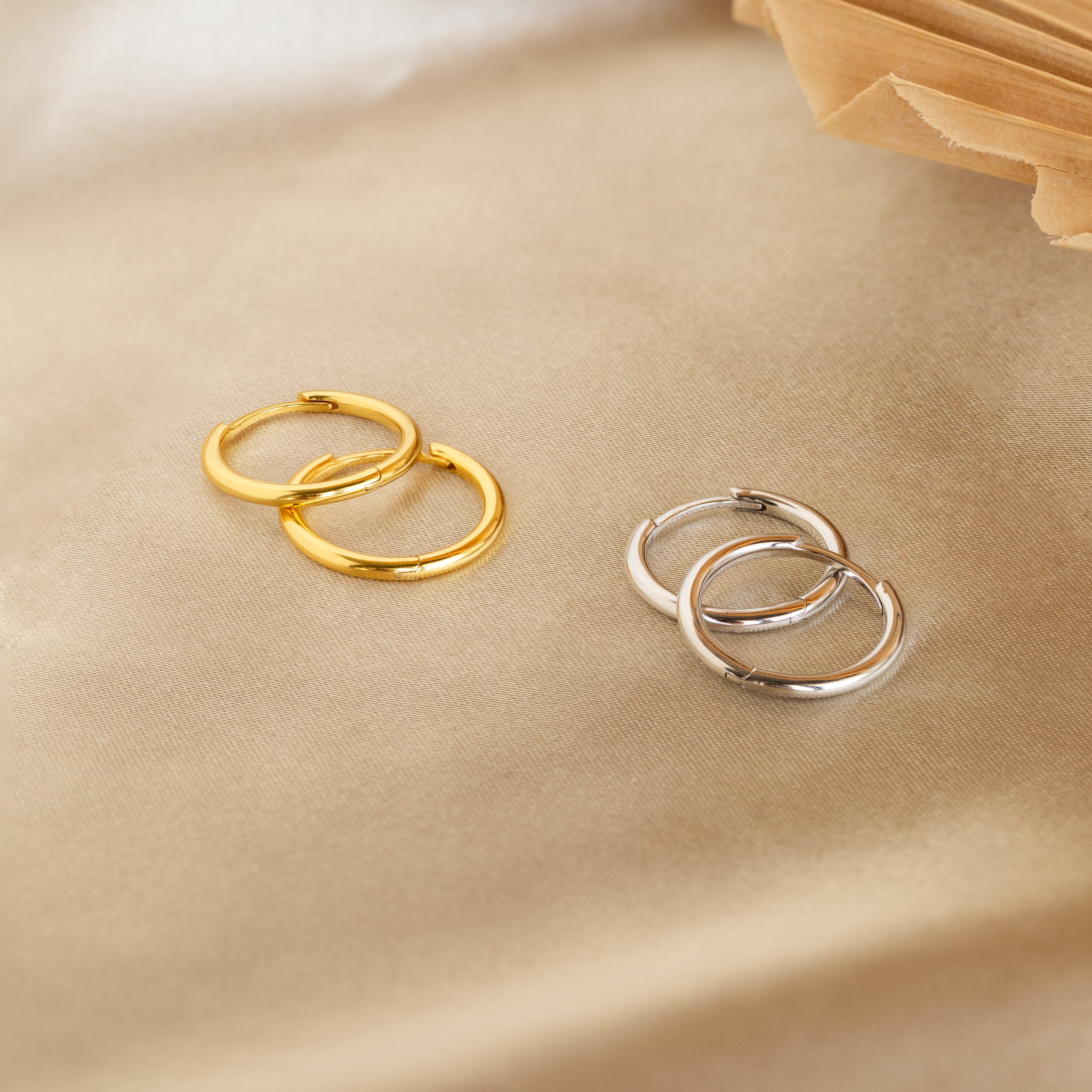 Two pairs of Medium Bold Hoops, one gold and one silver, rest on beige satin—lightweight earrings perfect for everyday jewelry style.