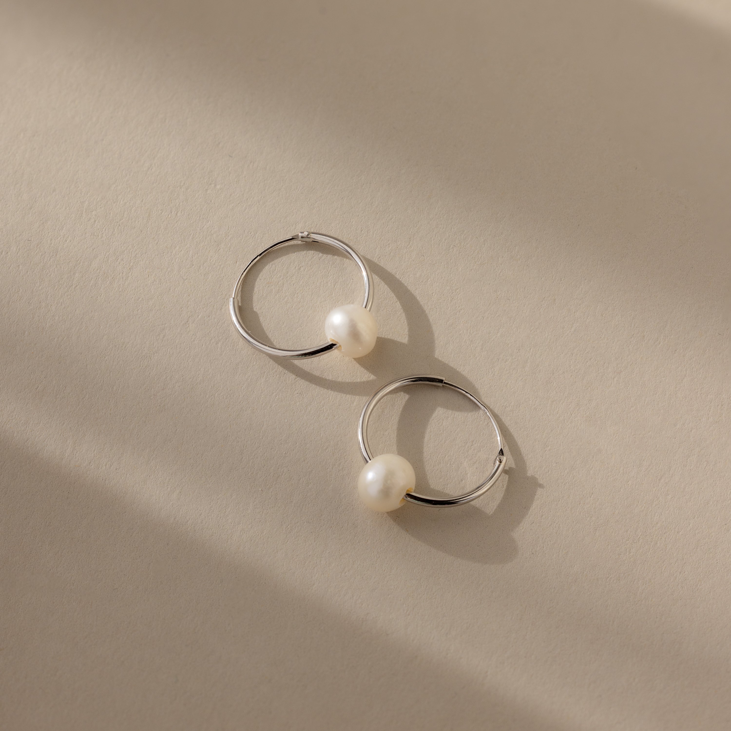 The Round Pearl Hoops in Sterling Silver, showcasing polished sterling silver hoops each adorned with a luminous pearl, are displayed on a beige surface with soft shadows.