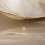 Close-up of a delicate silver chain necklace featuring a single round green jade bead, draped from a white shell dish.