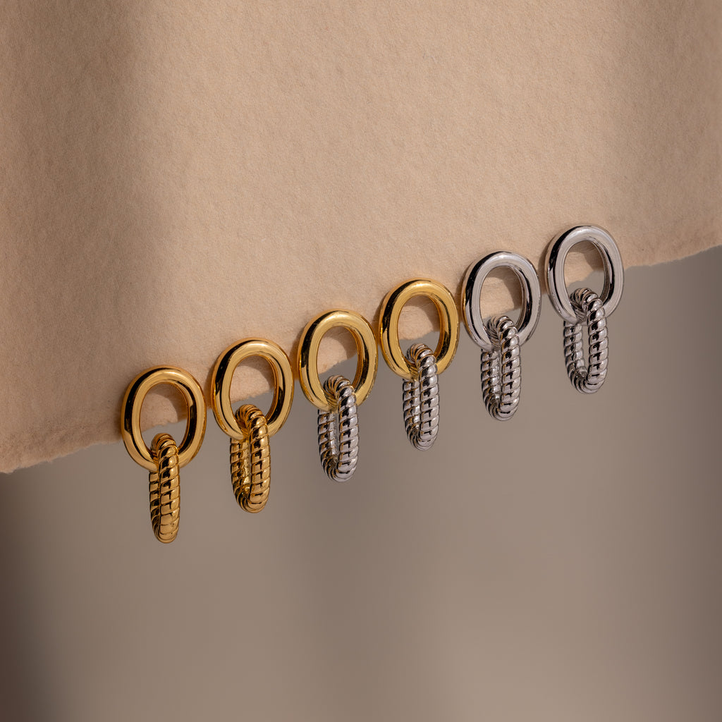 Six metallic rings, three gold and three silver, hang in a row from a beige fabric edge—mirroring the sculptural elegance of the Twisted Link Earrings.
