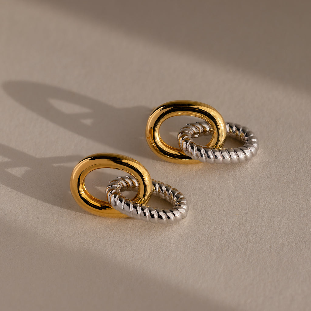The Twisted Link Earrings in Mixed Metal feature interlocking gold and silver textured designs, highlighting sculptural elegance against a neutral background.