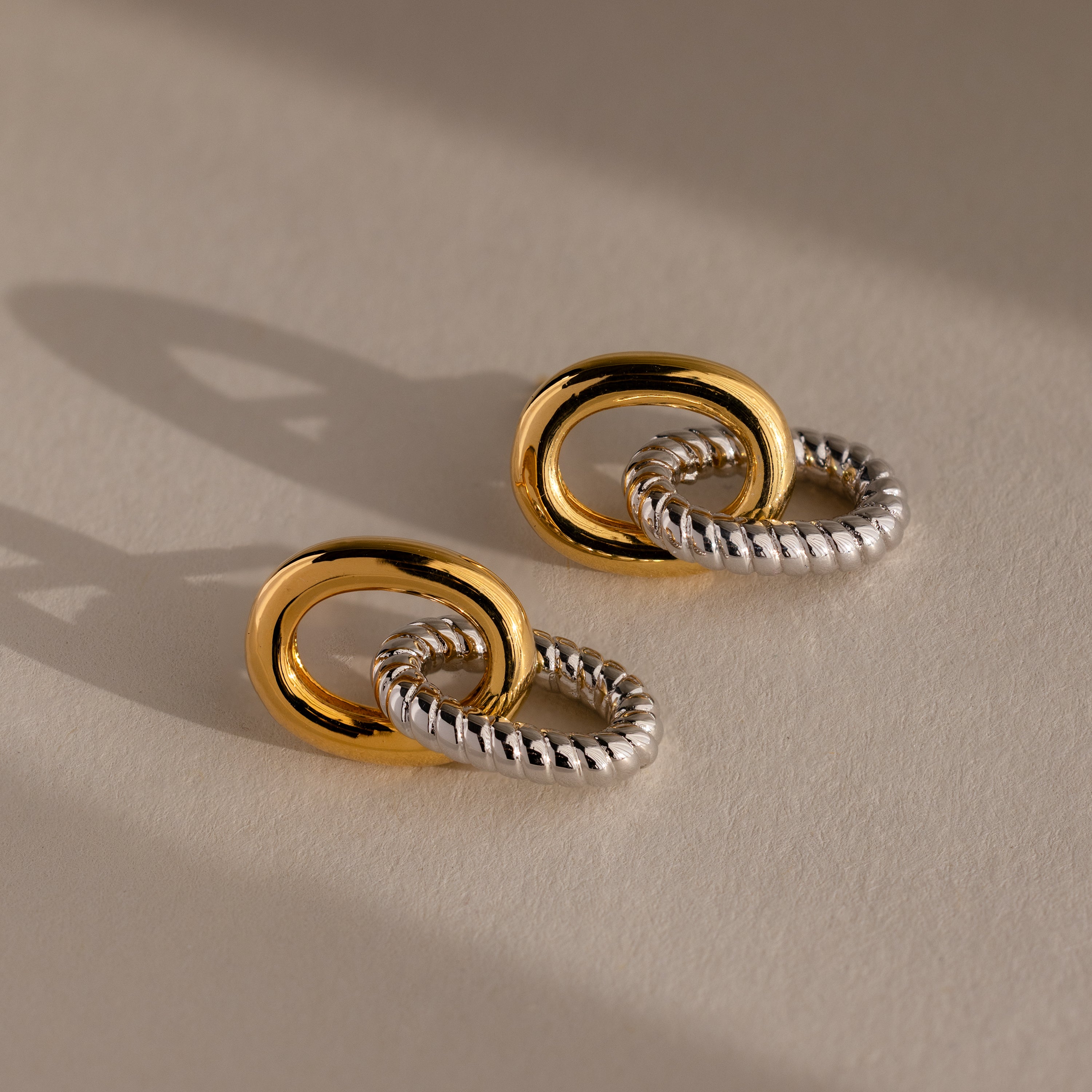 The Twisted Link Earrings in Mixed Metal feature interlocking gold and silver textured designs, highlighting sculptural elegance against a neutral background.