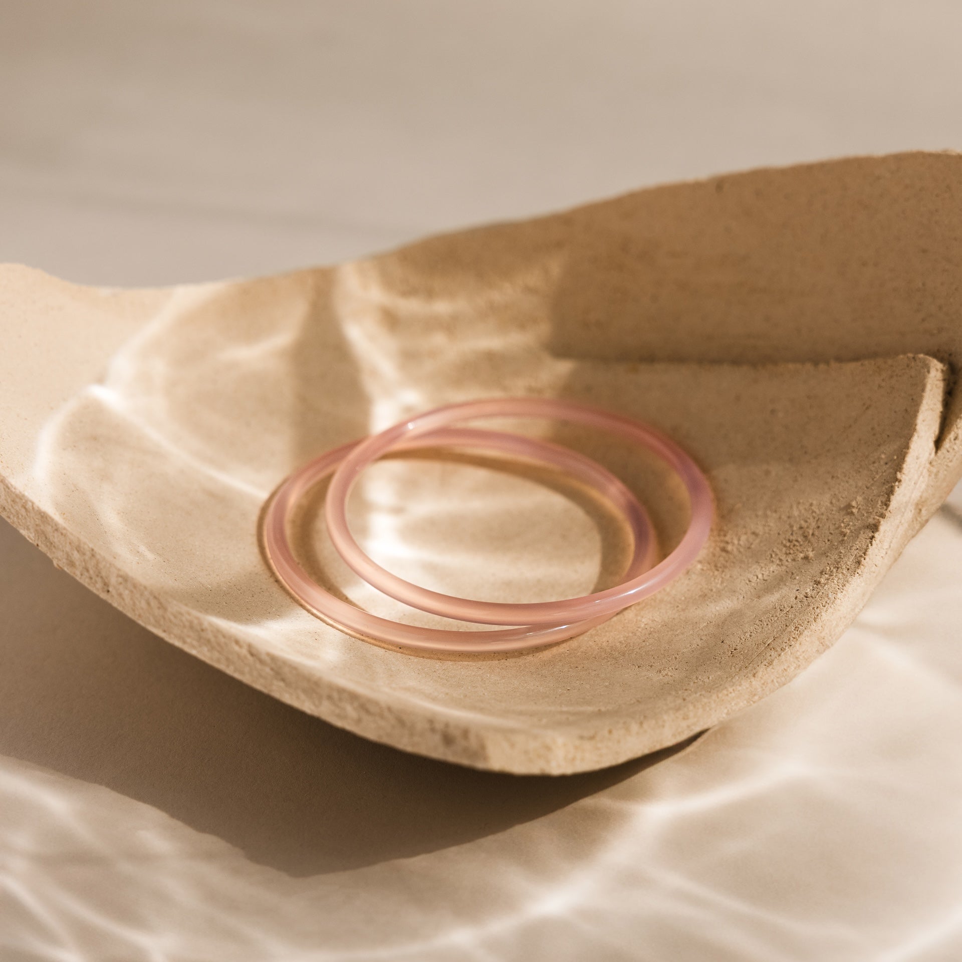 The Thin Jade Bangle Set, with a glass-like finish in pale pink, rests on a textured beige ceramic dish, catching soft light reflections.