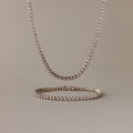 The Diamond Bezel Tennis Necklace & Bracelet Set in White Gold, displayed on a beige background, makes an elegant and unforgettable anniversary gift.
