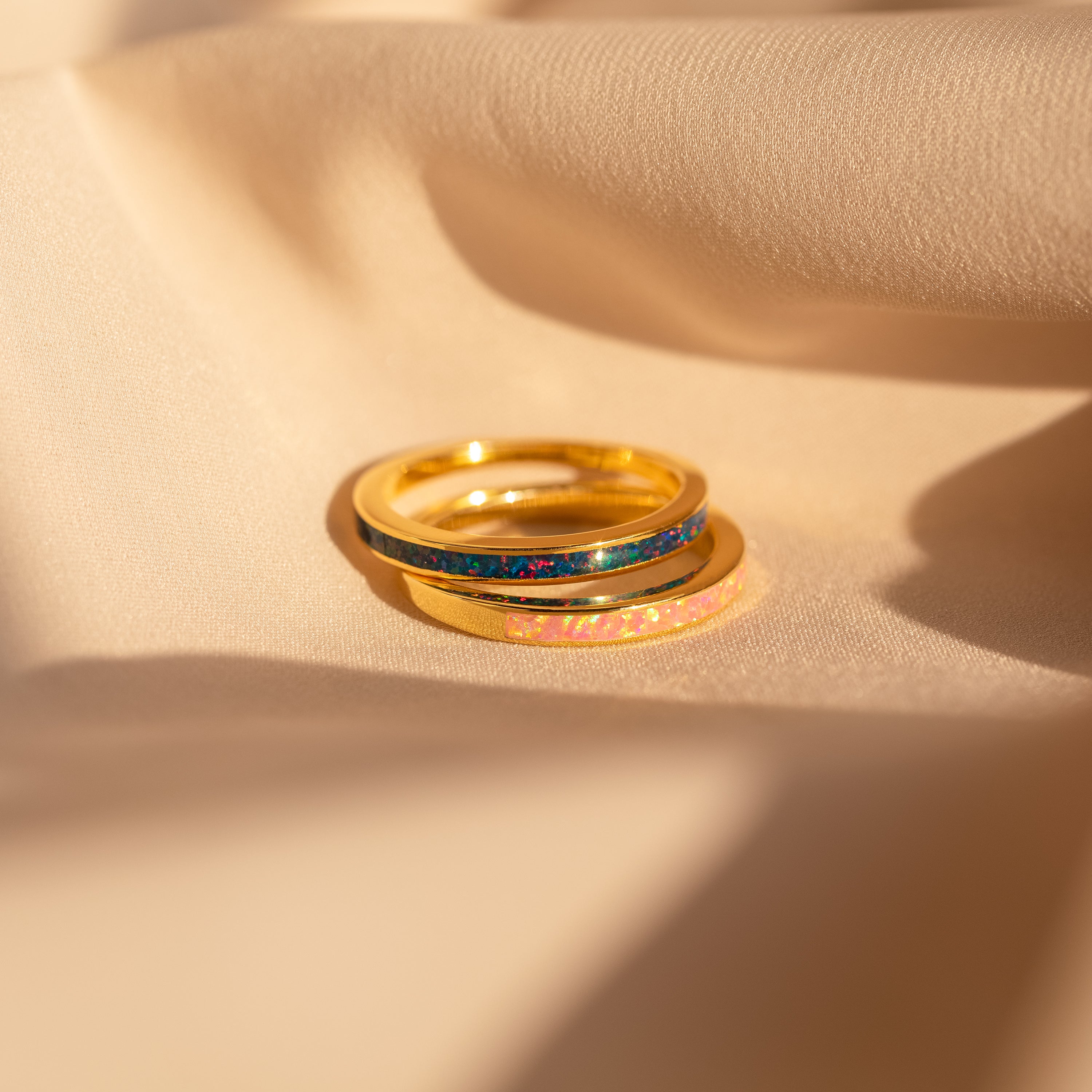 The Opal Inlay Rings Set, with Indigo Blue and Pink Flamingo accents, is stacked on beige fabric and glows gently in the sunlight.