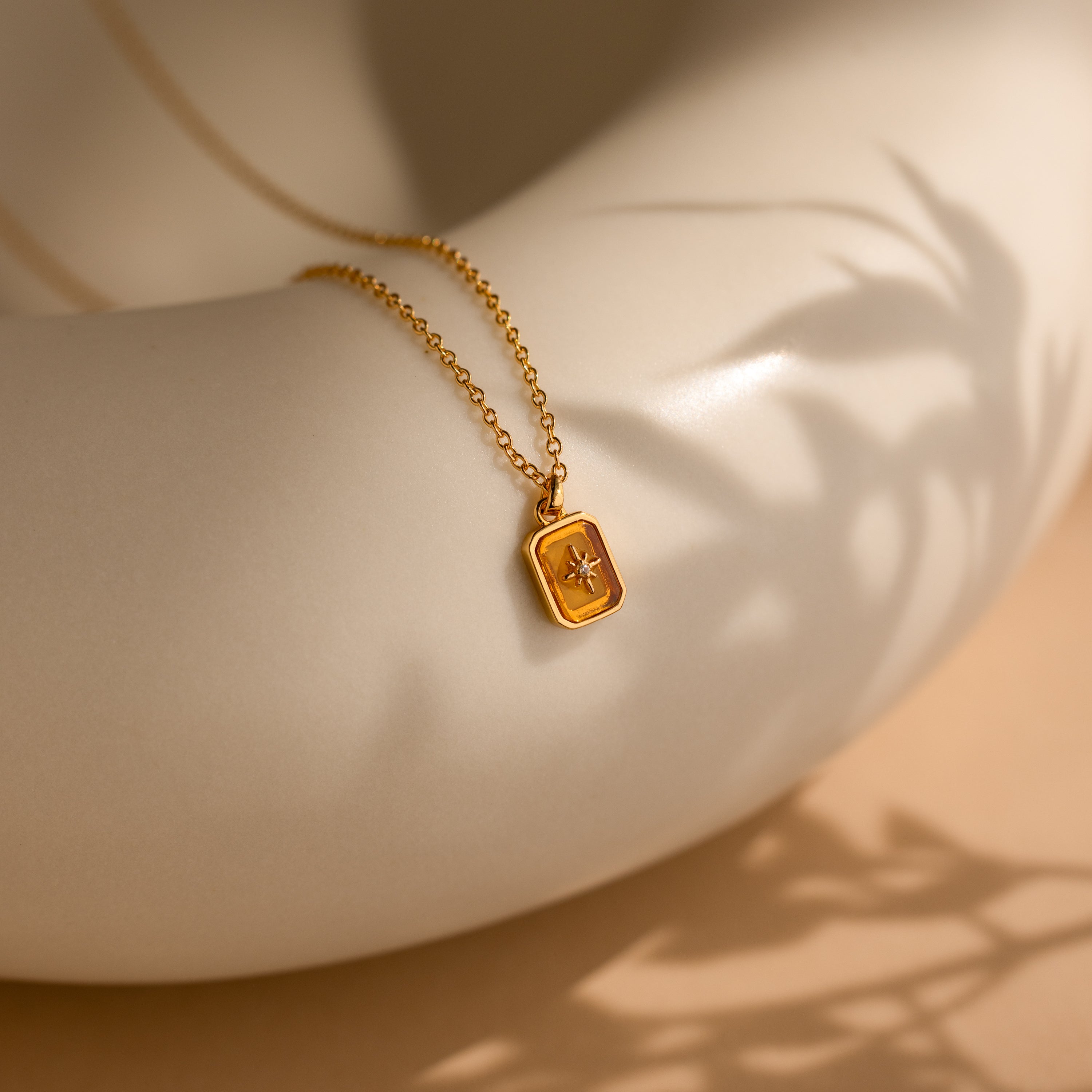The North Star Birthstone Necklace in Topaz (November) features a rectangular pendant with a diamond-set North Star, elegantly draped over a smooth, white curved surface.