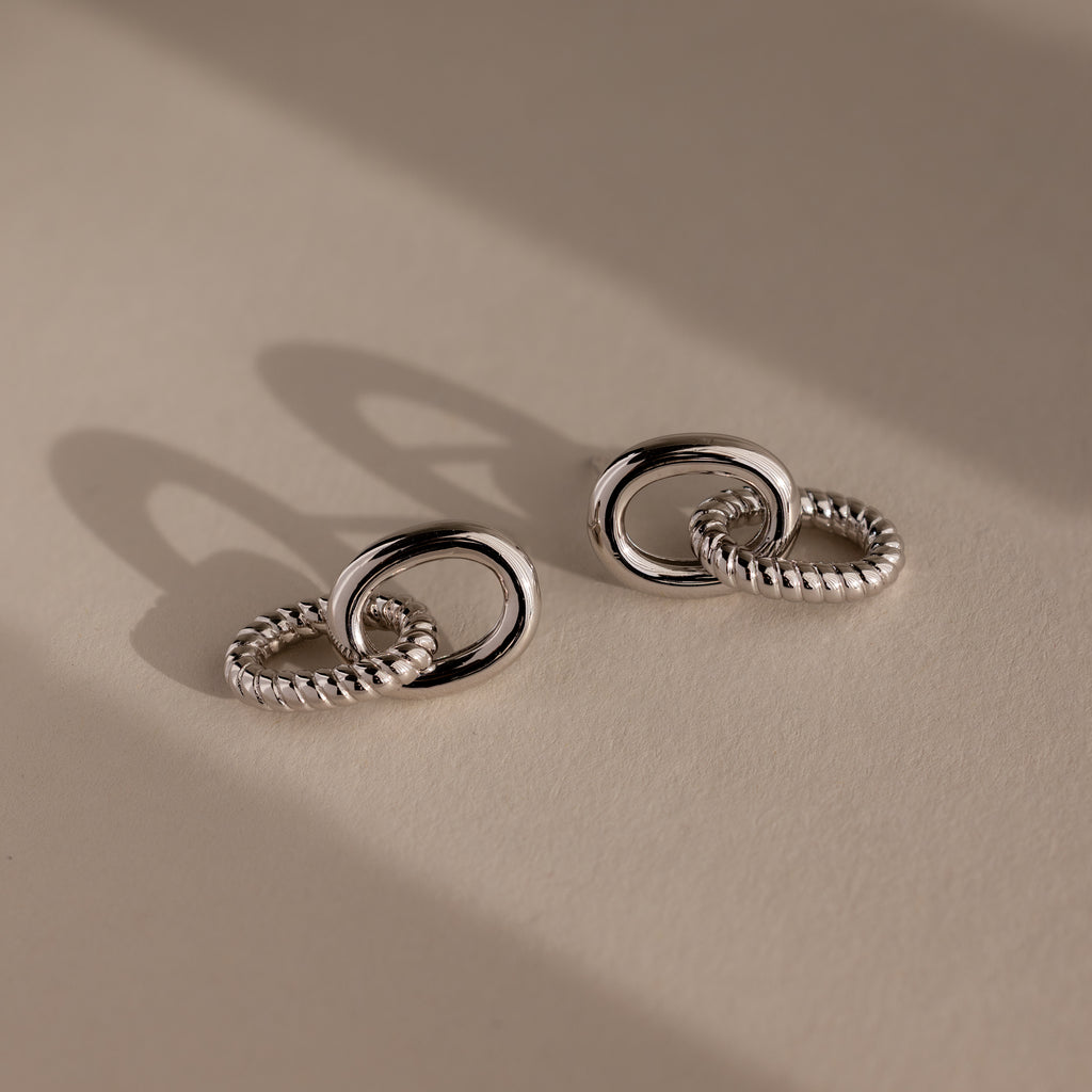 Two pairs of Twisted Link Earrings with silver intertwined circles cast soft shadows on a neutral surface—striking pieces of modern jewelry.