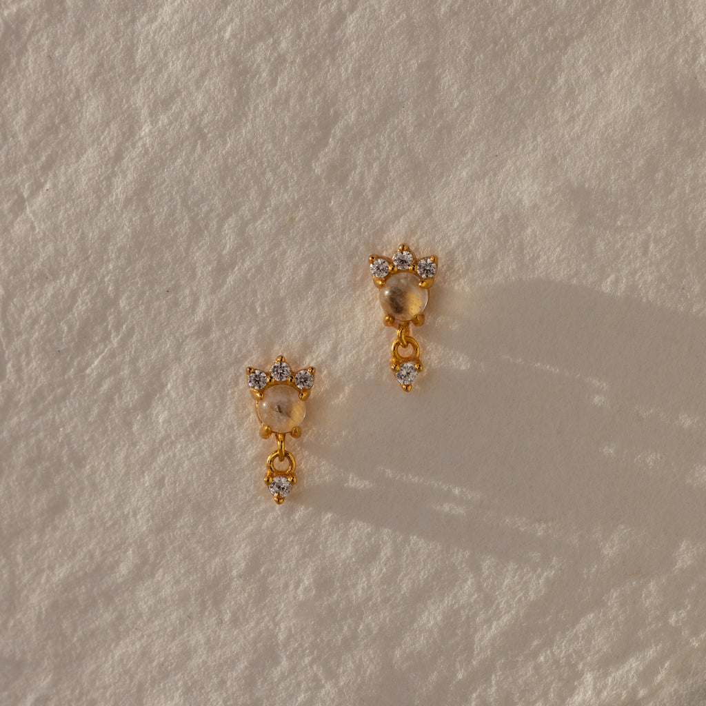 The Halo Moonstone Drop Studs in 18K Gold radiate royal elegance, their round stones shimmering against a textured cream backdrop.