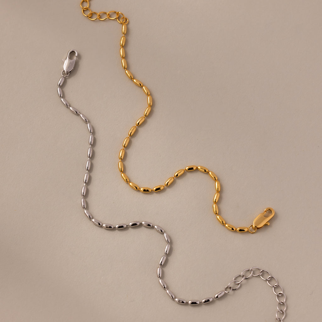 The Oval Beaded Bracelet features polished gold and silver chains with lobster clasps, displayed on a beige surface—a modern classic jewelry piece.
