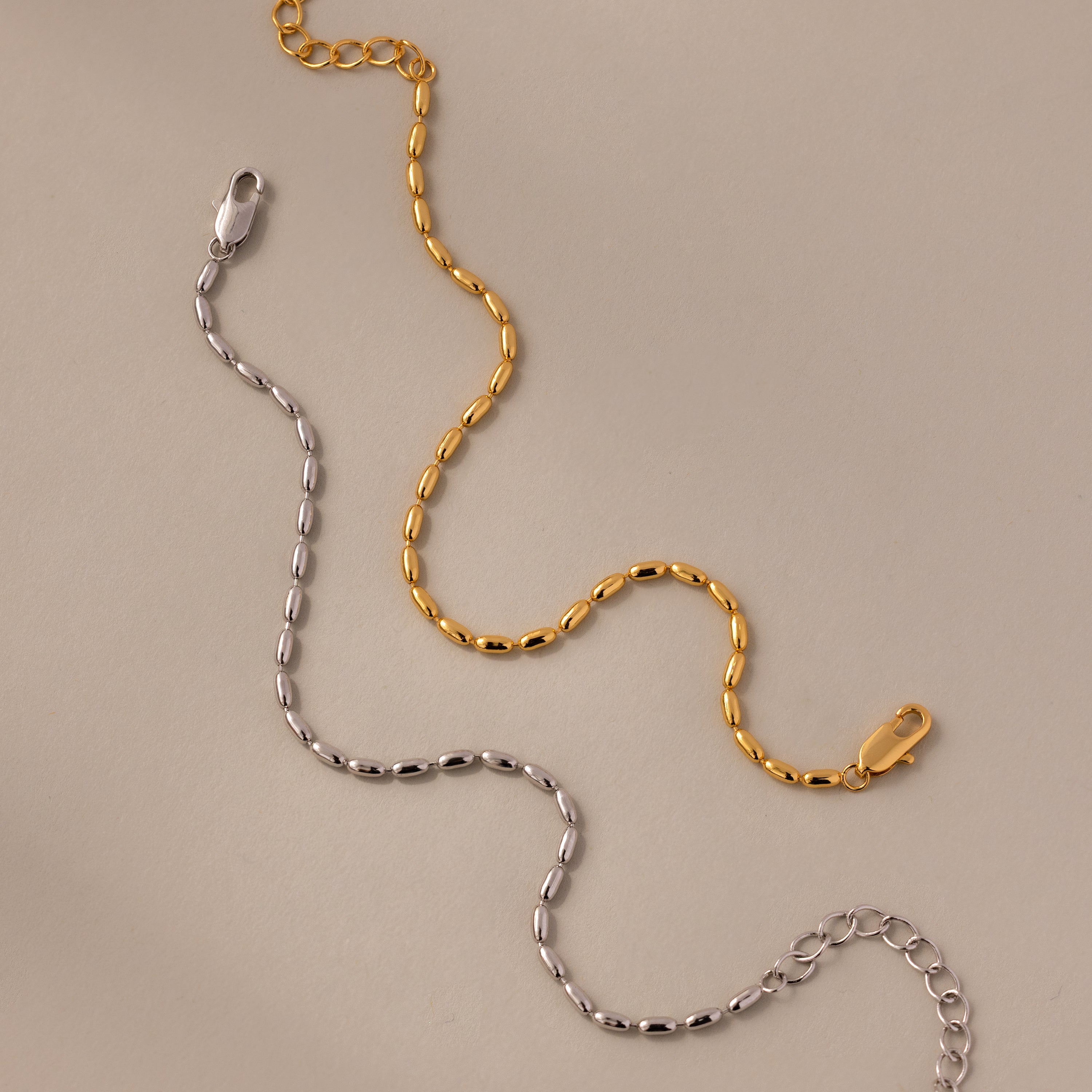 The Oval Beaded Bracelet features polished gold and silver chains with lobster clasps, displayed on a beige surface—a modern classic jewelry piece.