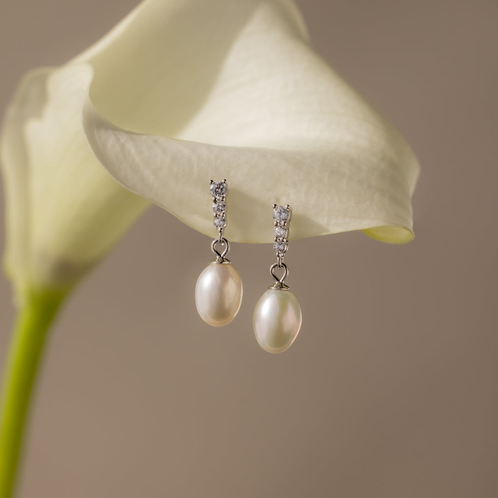 The Diamond Pearl Droplet Earrings in Sterling Silver rest gracefully on a white calla lily, their elegant design and sparkling studs capturing refined beauty.