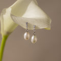 The Diamond Pearl Droplet Earrings in Sterling Silver rest gracefully on a white calla lily, their elegant design and sparkling studs capturing refined beauty.