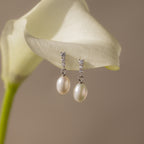 The Diamond Pearl Droplet Earrings in Sterling Silver rest gracefully on a white calla lily, their elegant design and sparkling studs capturing refined beauty.
