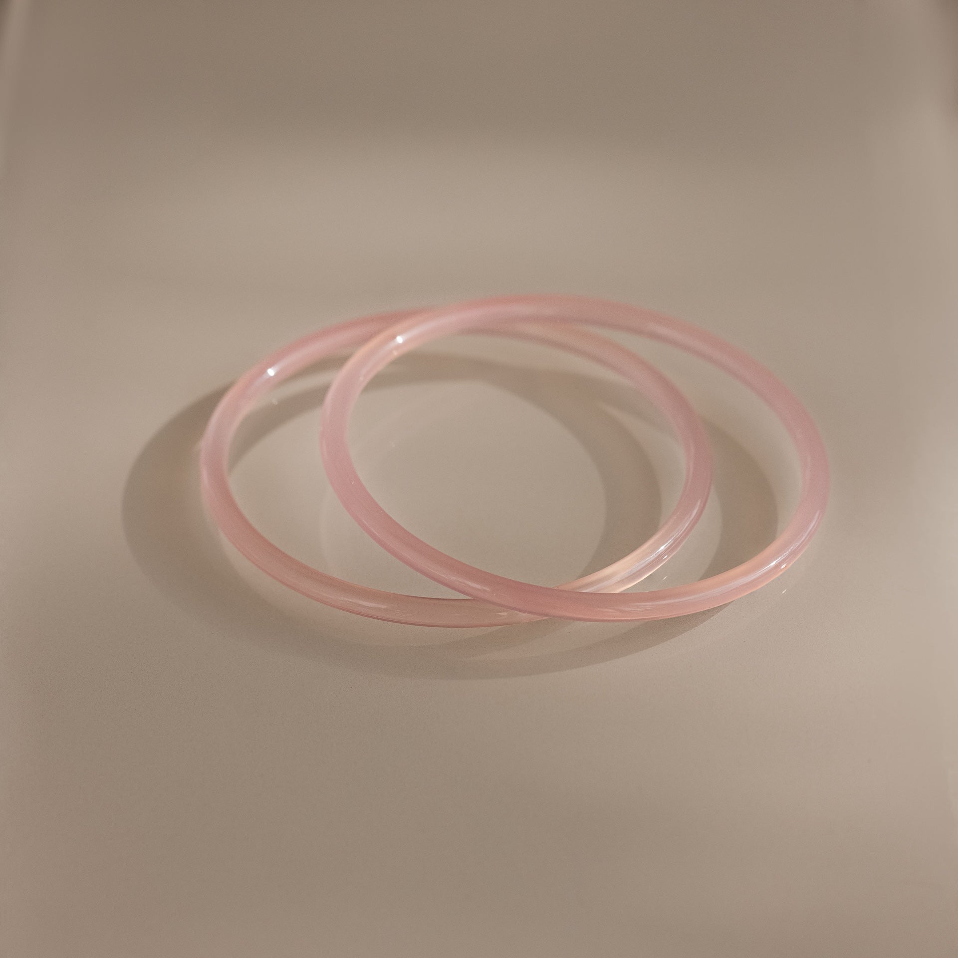 The Thin Pink Jade Bangle Set features two slim, light pink bangles that overlap elegantly on a cream surface—ideal for creating a delicate layered bracelet look.
