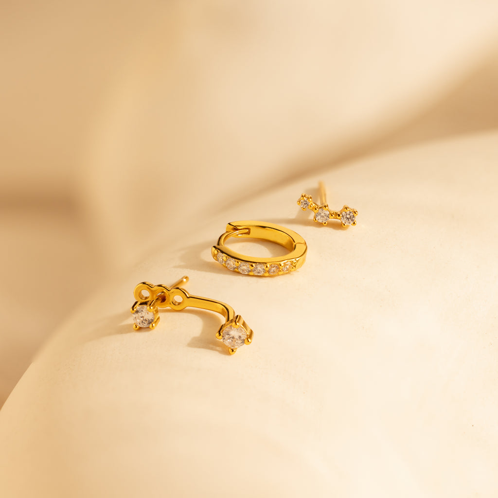 Three pieces of gold jewelry, including a Diamond Earrings Set in 18K Gold with sparkling stones, are displayed on a soft, cream-colored surface.
