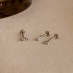 The Tiny Diamond Flatback Studs Set in Sterling Silver features three dainty earrings with clear gemstones, displayed on a light beige surface.