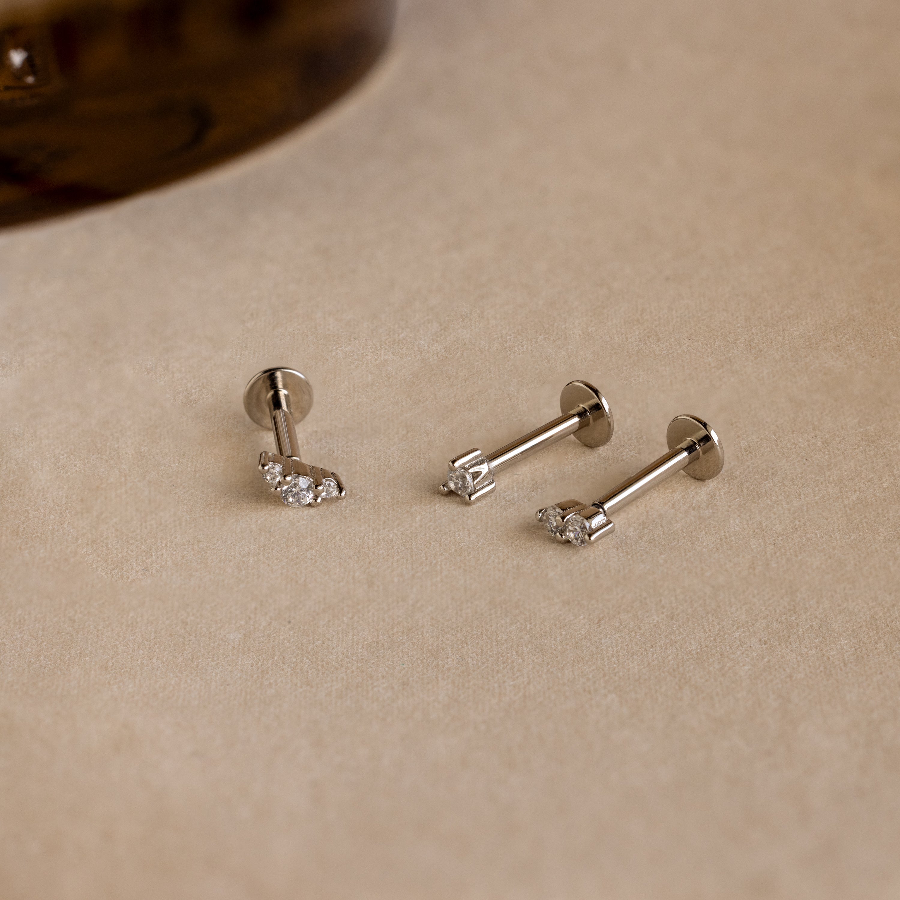 The Tiny Diamond Flatback Studs Set in Sterling Silver features three dainty earrings with clear gemstones, displayed on a light beige surface.