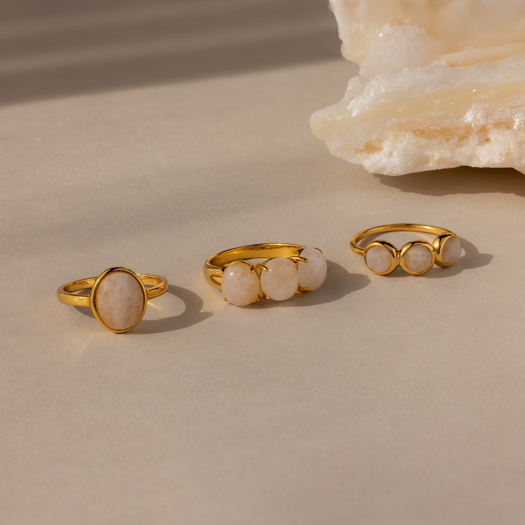 Three gold statement rings with white stones, including the eye-catching Cosmic Moonstone Ring, are displayed on a beige surface beside a textured white rock—perfect for any boho jewelry collection.